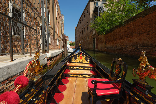Venice, Italy - August 23, 2026: Point of view from inside an ornate Venetian gondola with red velvet armchairs and gilded carvings cruising down a sunny narrow water canal alongside aged brick.