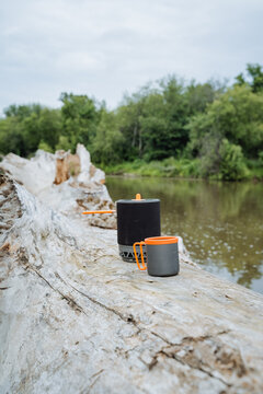 Outdoor camping cookware and insulated mugs rest on pale driftwood beside still river, with green forest shoreline, soft overcast light, orange handle accents, and rustic travel gear ready for meal