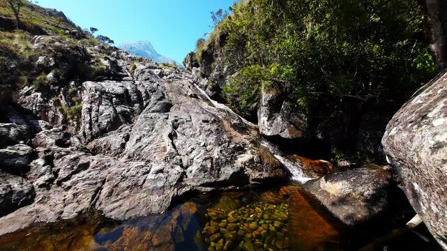 A gentle waterfall trickles down sloped rock into a clear mountain pool on Mulanje Mountain, with rugged vegetation and a distant peak on the horizon.