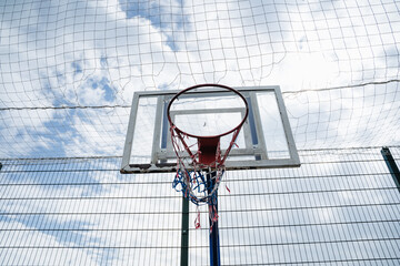 Low angle view of worn outdoor basketball hoop showing chipped backboard, rusted rim, and frayed red white blue net in front of tall protective fence and cloudy sky, creating gritty urban sports scene © Aleksey