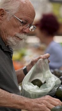 Elderly man selects and examines avocados while grocery shopping in a local store. He carefully checks the quality of the fruit.