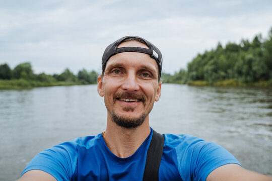 Close-up selfie shows smiling adult man wearing backward cap and blue shirt during river paddling, with calm water, tree-lined shore, soft overcast light, and relaxed outdoor recreation mood
