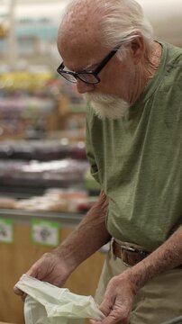 An elderly man picks items from shelves and puts them in a bag at a grocery store in the afternoon. He focuses on his task.