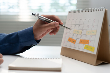 Business professional pointing at a desk calendar with scheduled notes while planning appointments, deadlines, meetings and tasks for organized time management and workplace productivity. © Father_Studio