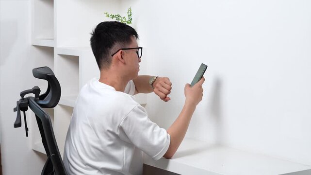 Asian young man sitting at white desk smartphone, checking watch, side view mockup with copy space