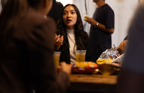 Asian woman engaging in conversation at social gathering. Colleagues enjoying an after-work party, socializing while sitting at a table with drinks and snacks.
