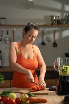 Young woman preparing fresh smoothie cutting carrot in kitchen. healthy meal, vegan diet and detox