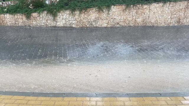Heavy rain splashes onto a paved driveway as muddy runoff flows beside a curb and landscaped garden beds. Water covers the paving stones during the downpour.