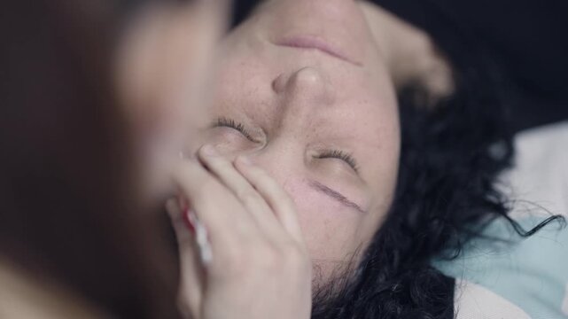A woman is applying makeup on eyebrows to another woman's face lying on a couch
