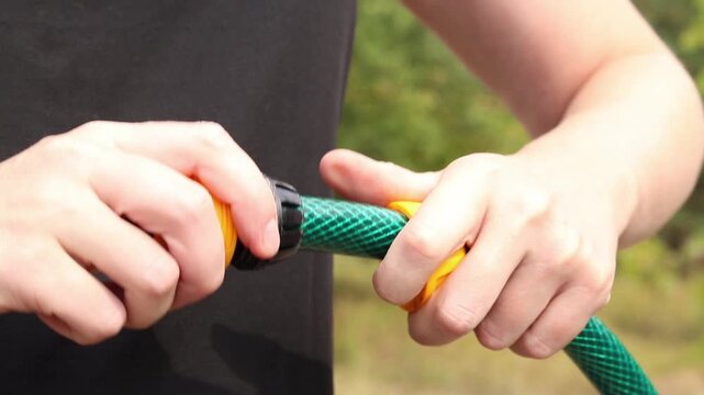Close-up of hands tightening yellow plastic connector (quick release) on green garden hose on blurred background of green leaves in garden. Garden care, preparing watering equipment