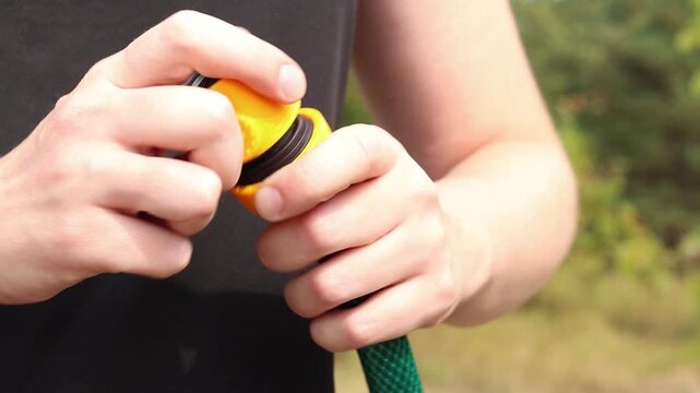 Close-up of hands tightening yellow plastic connector (quick release) on green garden hose on blurred background of green leaves in garden. Garden care, preparing watering equipment