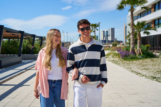Young couple enjoying a sunny day stroll, linking arms on a modern walkway.