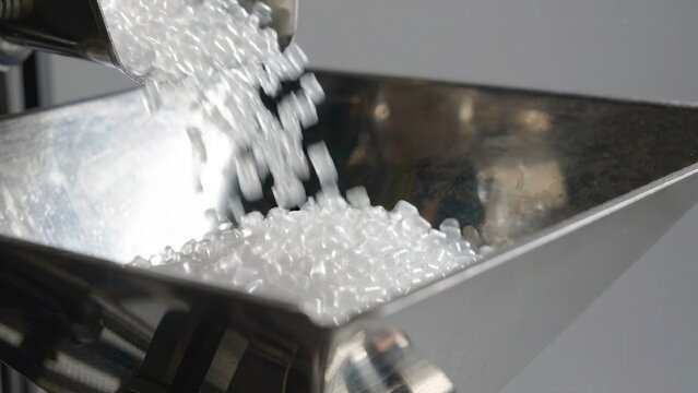White plastic pellets continuously flow from a metal hopper into a stainless steel container, showcasing the manufacturing process in a clean industrial setting