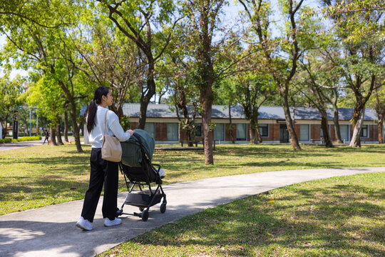 Asian mother pushing stroller on sunlit park pathway