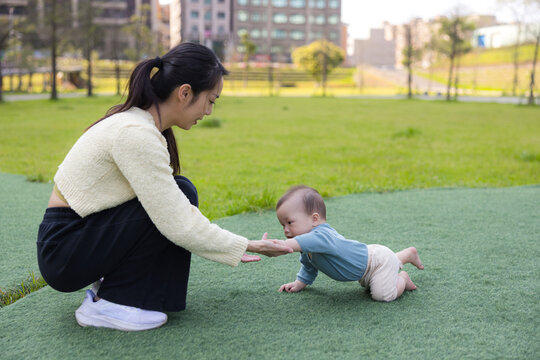 Mother helping baby crawl on grass in city park