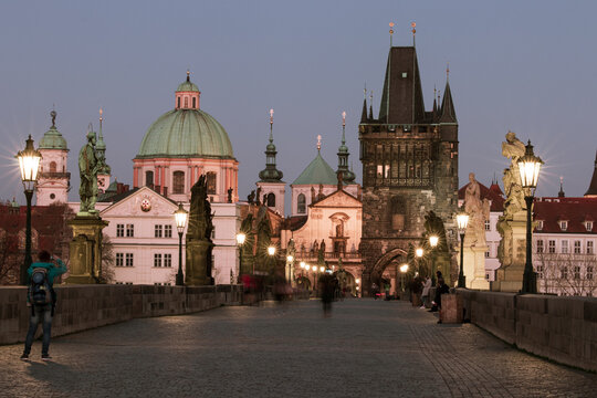 View from Charles Bridge toward the Old Town Bridge Tower before sunrise, with historic churches, statues and glowing street lamps in Prague, Czech Republic.