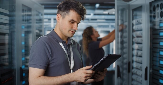 Professional IT Specialist Operates Tablet Computer to Monitor System Status while Female Colleague Inspects Server Rack Hardware. Team Collaboration Ensures Smooth Cloud Infrastructure Performance.