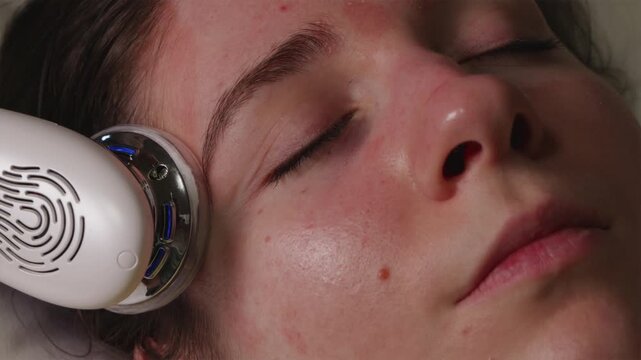 Close up of young woman receiving facial cleansing with blue LED light skincare device