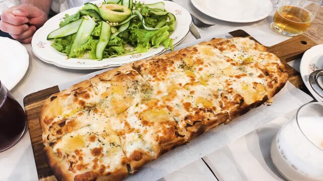 Delicious rectangular pizza bakes with four cheeses on wooden serving tray. Hot rectangular pizza topped with herbs serves for group dinner in restaurant with drinks on checked tablecloth.