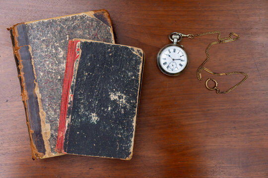 Old vintage books with worn covers and antique pocket watch on wooden table. Nostalgic still life of time and reading. Retro memory, history and antiques collection concept.