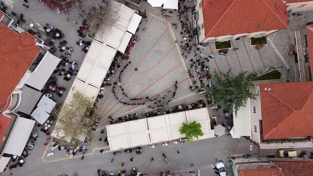 Aerial view of traditional folk dance spiraling through village square, Deskati, Greece. Dancers form a winding line amid red-roofed buildings.