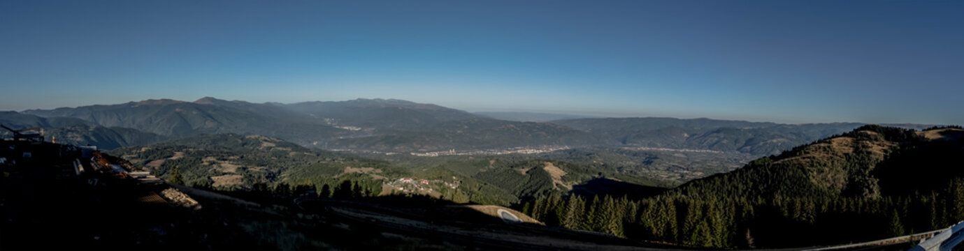 A panoramic view from the Par&acirc;ng Mountains towards the Jiul Valley, showcasing vast rolling hills, dense forests, and distant settlements under a clear blue sky.