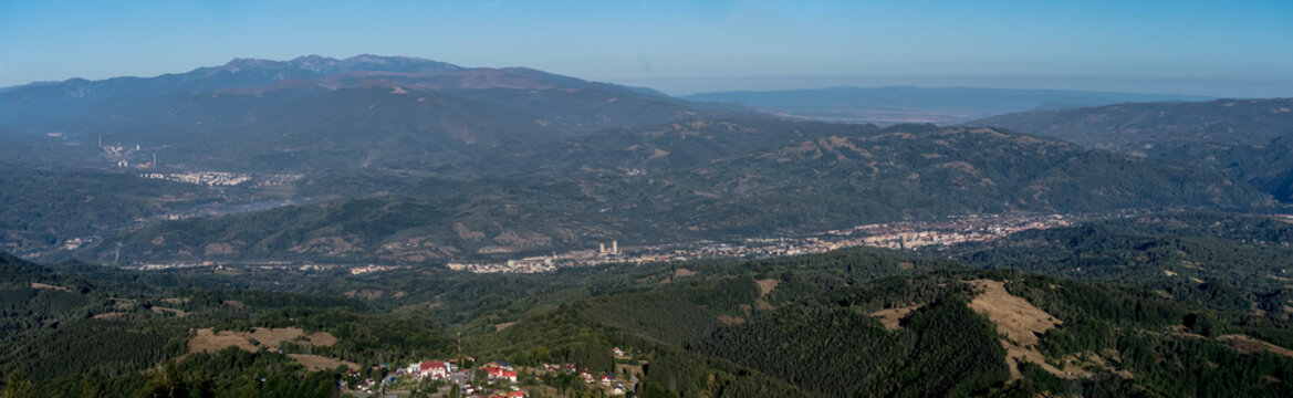 A panoramic view from the Par&acirc;ng Mountains towards the Jiul Valley, showcasing vast rolling hills, dense forests, and distant settlements under a clear blue sky.