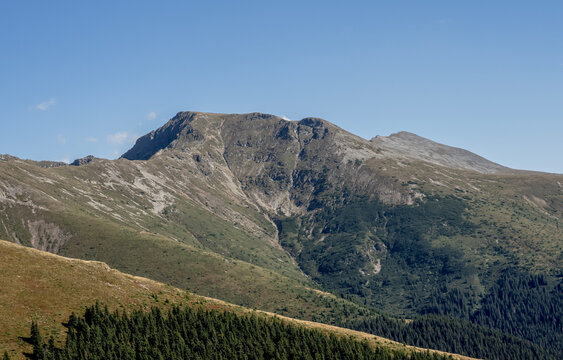 A scenic view of C&acirc;rja Peak (V&acirc;rful C&acirc;rja) and alpine slopes in the Par&acirc;ng Mountains, Romania, under a clear blue sky.