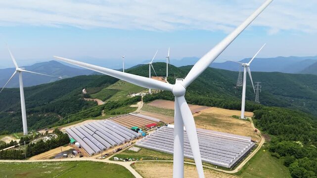 Aerial drone zooms toward close up of rotating wind turbine blades with vinyl greenhouses and farmland visible on mountain ridge below