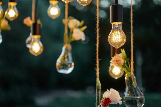 Warm glowing filament bulbs hang beside small glass vases filled with delicate flowers, creating a romantic, intimate outdoor wedding or party decoration with vintage ambiance and soft bokeh.