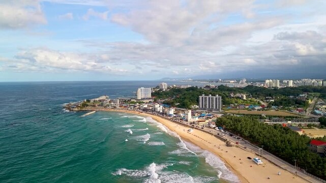 Drone parallax shot reveals Jumunjin Beach with rolling waves, sandy shoreline and waterfront city buildings under stormy clouds in Gangneung