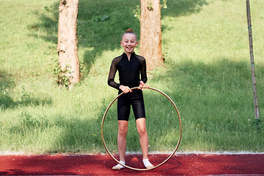 A cheerful young girl gymnast holding a hoop, standing on a path surrounded by lush greenery.