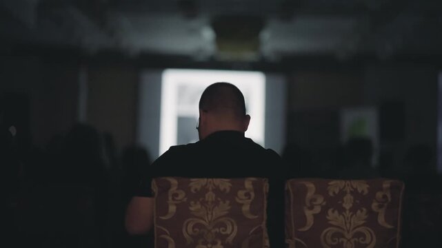 A man sits in a chair in a room full of people at business meeting summit. The manis looking at videobim presentation. The room is filled with people sitting in chairs