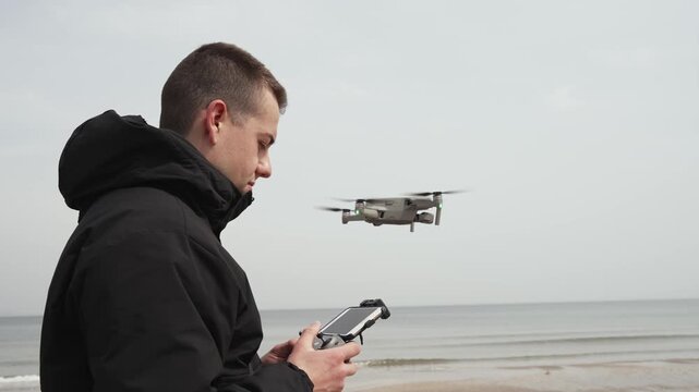 A man is holding a drone remote control. The scene is set on a beach, with the ocean in the background. The man is focused on the remote control