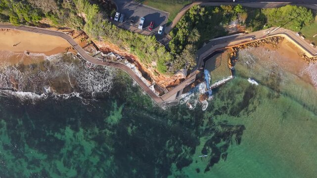 Terrigal Haven shoreline with rock pool, walkway and clear turquoise water, drone top down backdrop as waves crash on shores