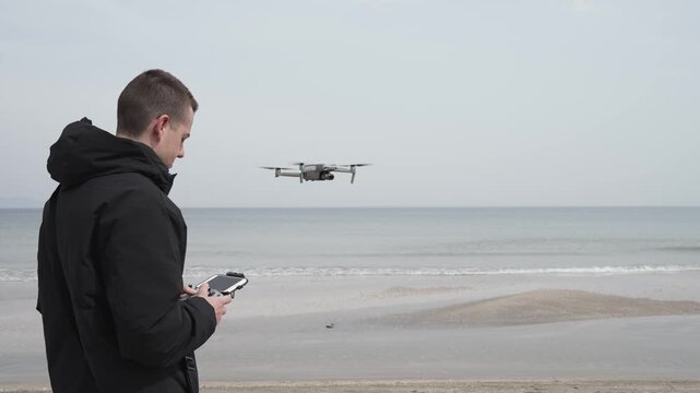 A man is holding a drone remote control. The scene is set on a beach, with the ocean in the background. The man is focused on the remote control
