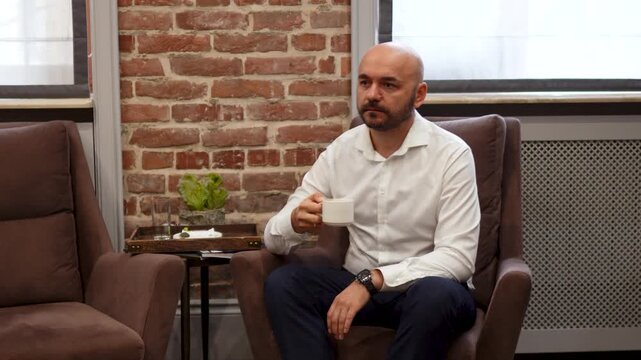 Adult man in white shirt sipping coffee while seated in brown armchair in brickwall lounge, then placing cup on round side table during calm indoor break moment