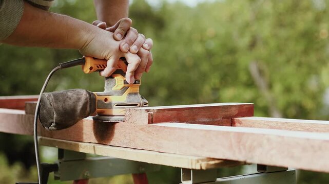 4K slow motion 60 fps footage of a male carpenter hands using an electric orbital sander machine to smooth a wooden beam outdoors during daytime.