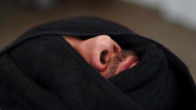 Closeup of male face wrapped in dark cloth showing nose, moustache, and slightly parted lips suggesting gentle breathing and resting state under soft indoor light