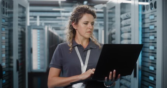 Focused Female Network Engineer Types on Laptop Computer While Standing Near Server Cabinets in Data Center. Hardware Configuration and Server Diagnostics Ensure Continuous Cloud Connectivity.