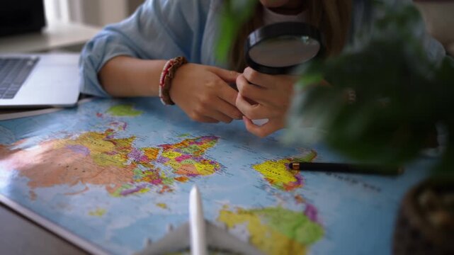 Curious teenage girl use magnifying glass to explore world map at home, study countries and destinations while discovering geography, learning about different cultures and developing interest in world