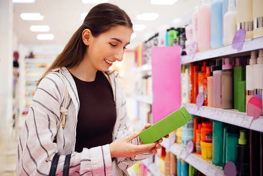 Young woman with long brown hair examines green product box while shopping in a beauty supply store, colorful bottles and containers visible on shelves
