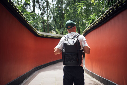 Male traveler wearing a black backpack walks through a narrow pathway lined with vibrant red walls and lush greenery in a serene outdoor setting