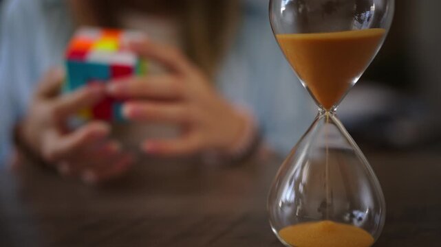 Hourglass with golden sand measuring time in foreground, with an out of focus young girl attempting to solve colorful puzzle cube in  background, symbolizing problem solving and patience