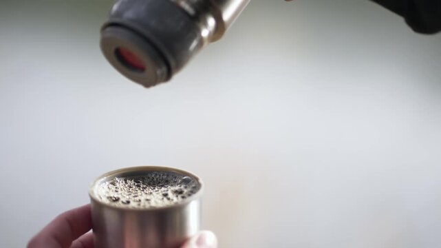 A person is pouring hot water into a thermos. The person is wearing a black jacket and he is outdoors