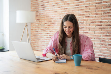 Young woman smiling, sitting at a wooden table and writing notes in a small spiral notebook while...