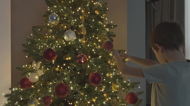 Boy decorating Christmas tree placing ornament among glowing lights. Warm festive moment. Suitable for family, lifestyle and holiday advertising.