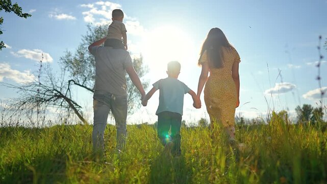 Happy family is walking in green park during summer. Parents and children are walking on grass at sunset. Father carries son on shoulders. Active family enjoys sunny summer walk together outdoors.