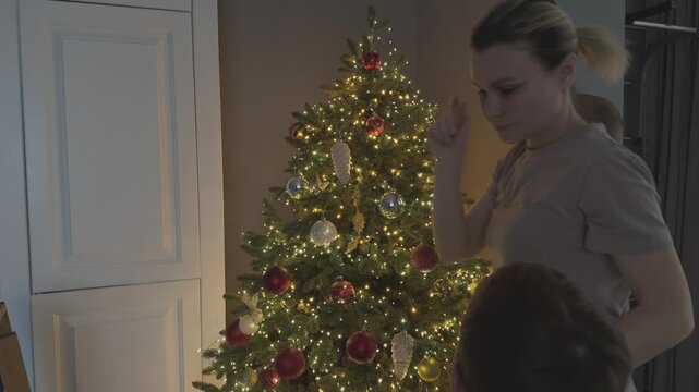 Mother helps child place decorations high on Christmas tree. Family bonding and festive preparation. Ideal for parenting, family and holiday storytelling.