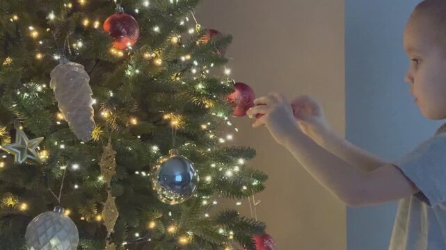  Small toddler decorating Christmas tree placing ornament carefully. Cute emotional family moment. Ideal for parenting content. 